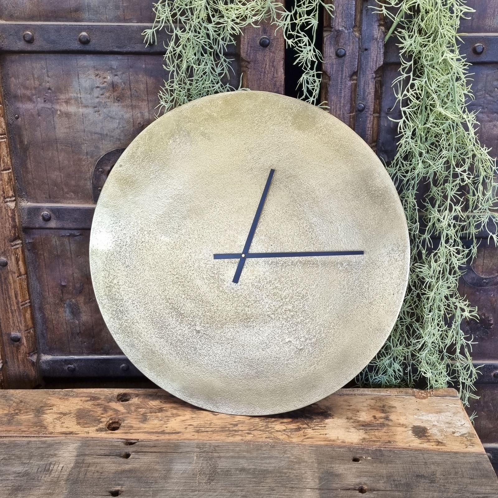 Eine Uhr Antik Bronze Klein mit schwarzen Zeigern, stehend vor einer alten Holztür mit grüner Hängepflanze - Benoa
