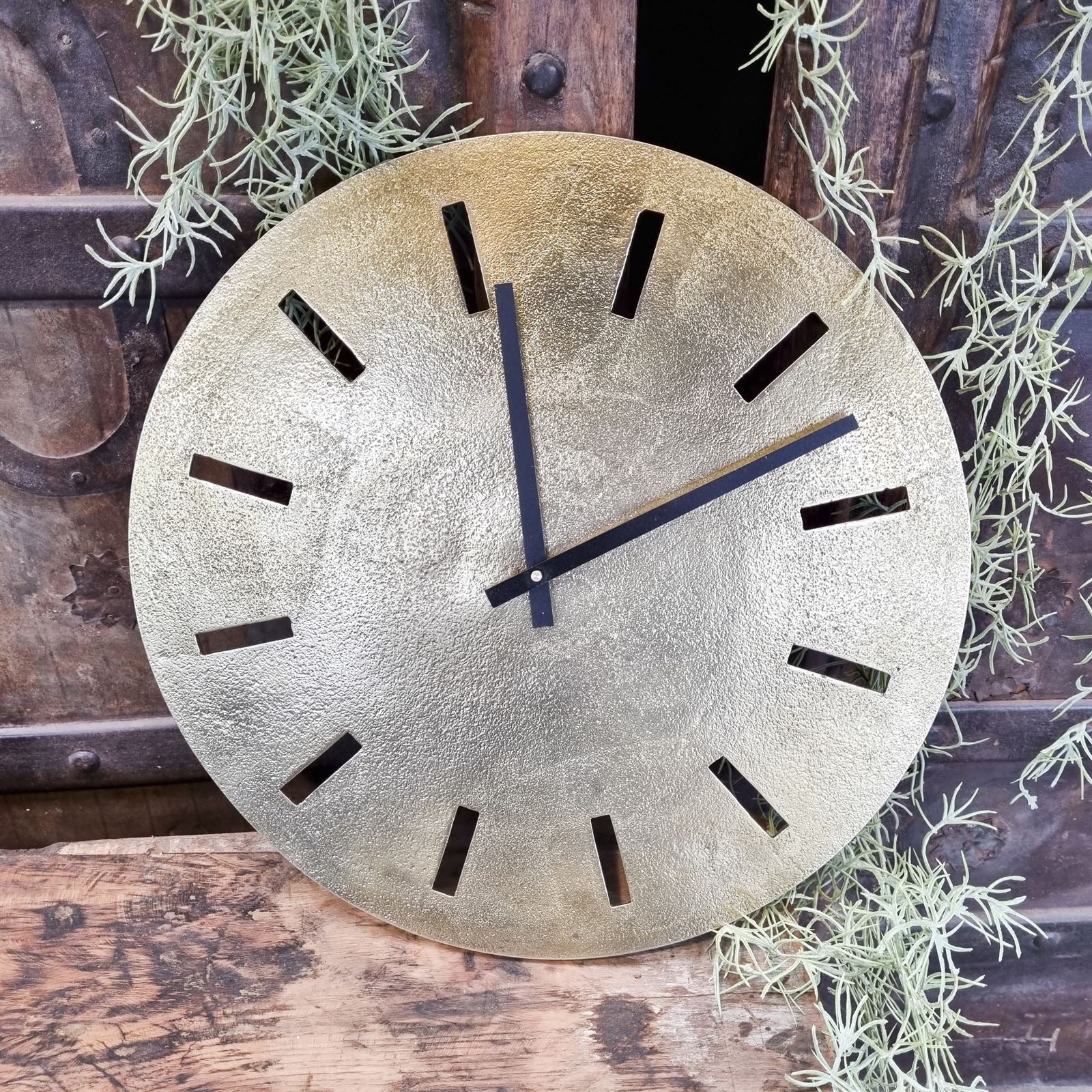 Große goldene Wanduhr mit schwarzen Zeigern und Stundenmarkierungen, stehend auf einem Holzuntergrund mit Pflanzen - Benoa