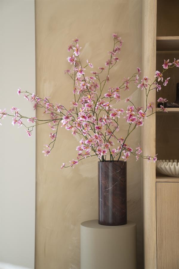 Die Vase Rosso mit rosa Orchideen steht auf einem beigen Sockel vor einer hellbraunen Wand - By-Boo