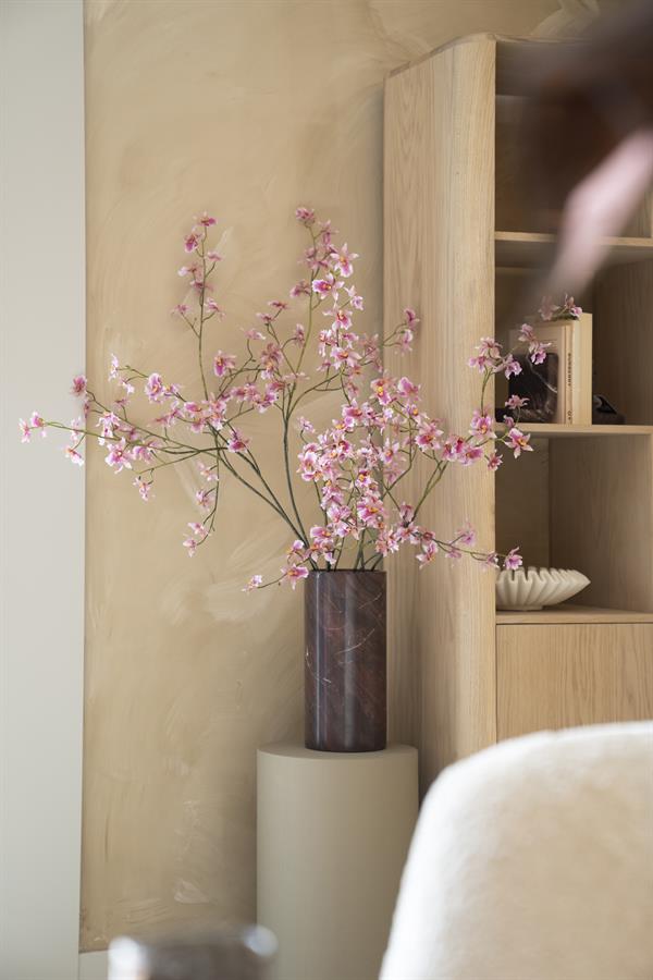 Die Vase Rosso auf einem beigefarbenen Sockel mit rosa Blüten, neben einem Holzschrank in einem Interieur - By-Boo