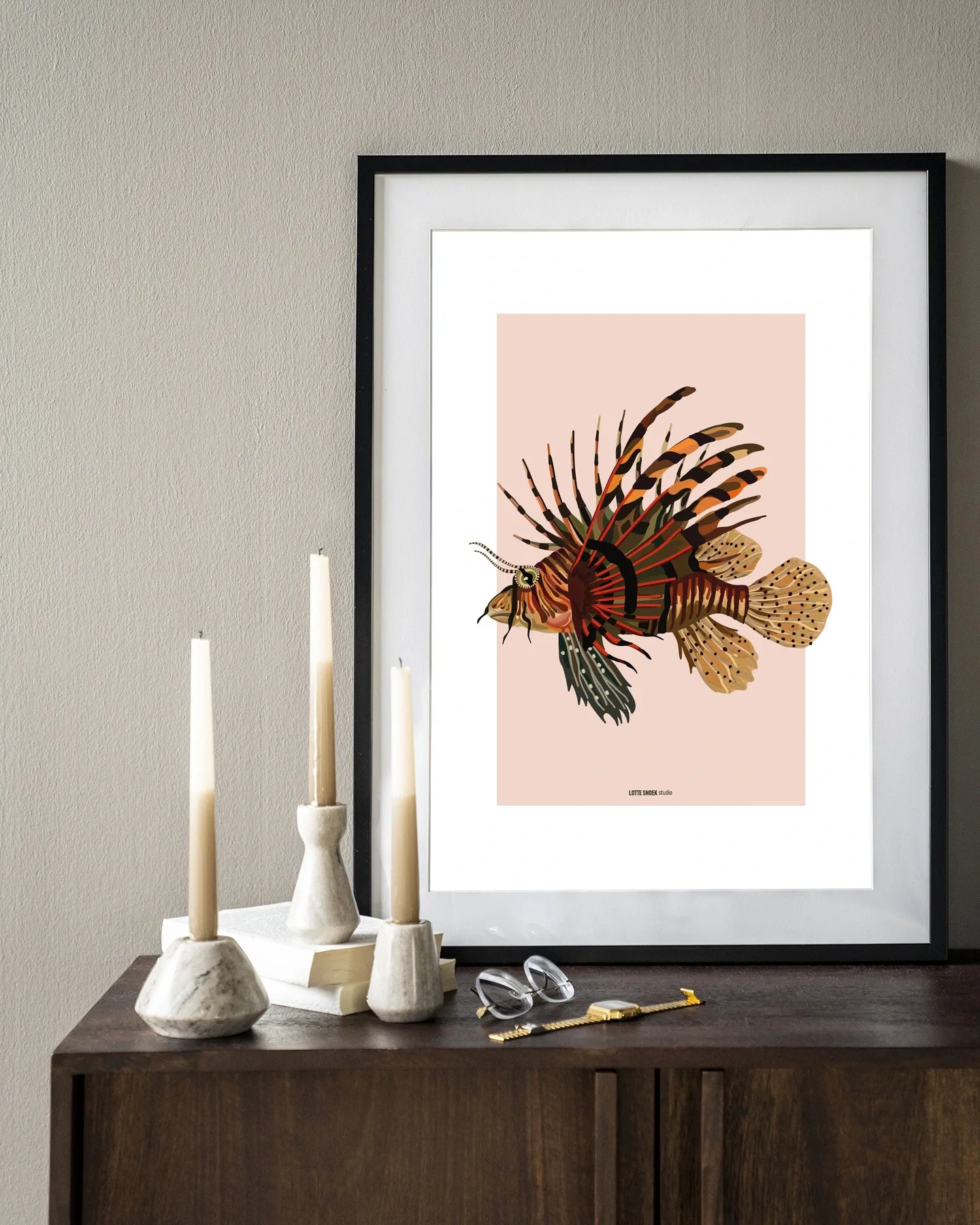 Een ingelijste Art print Lio - Urban Cotton met een afbeelding van een koraalduivel, staand op een houten dressoir met kaarsen en boeken