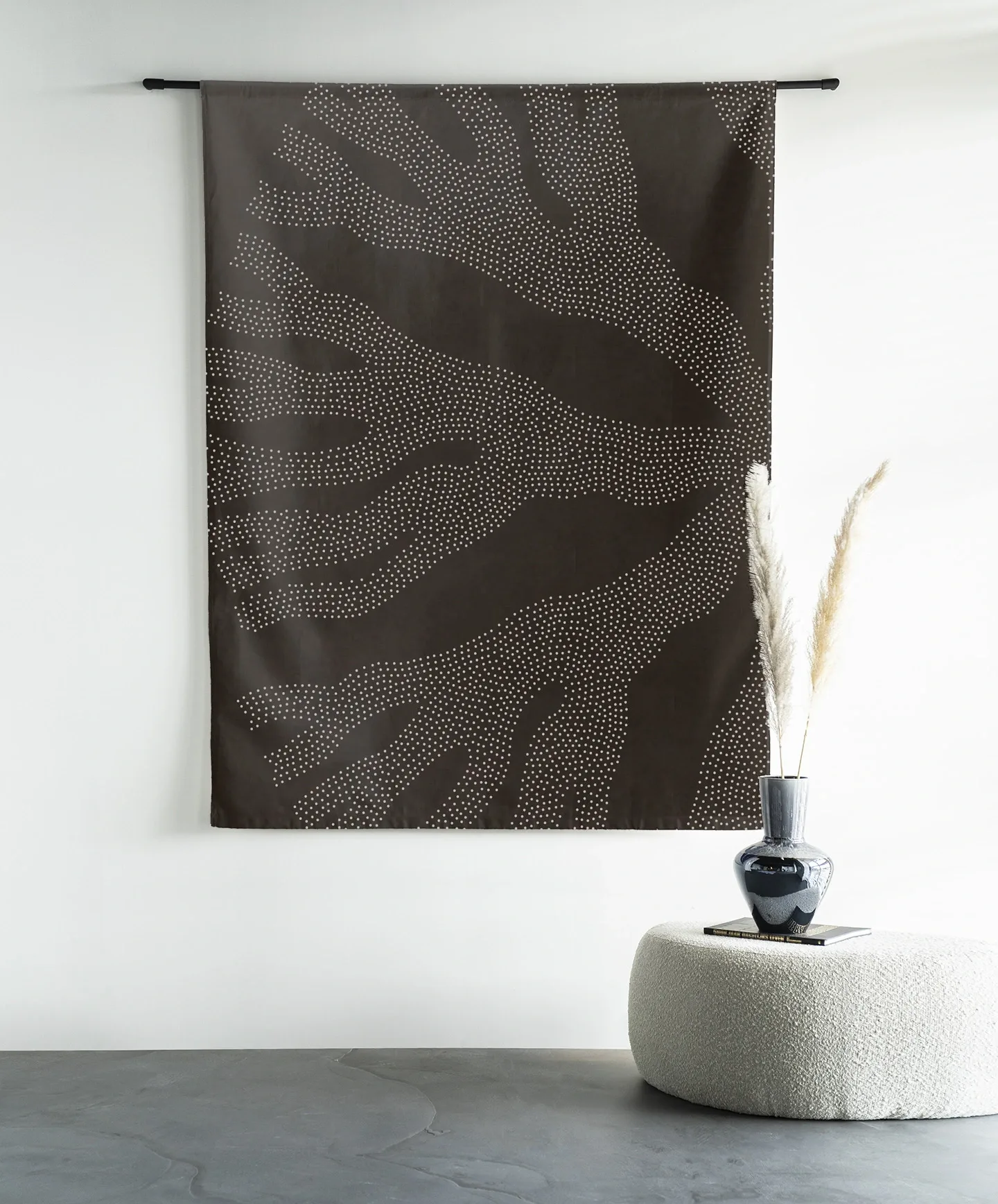 Wandteppich Circle Dark von Urban Cotton hängt an einer schwarzen Stange über einem weißen Pouf mit einer Vase und Pampasgras