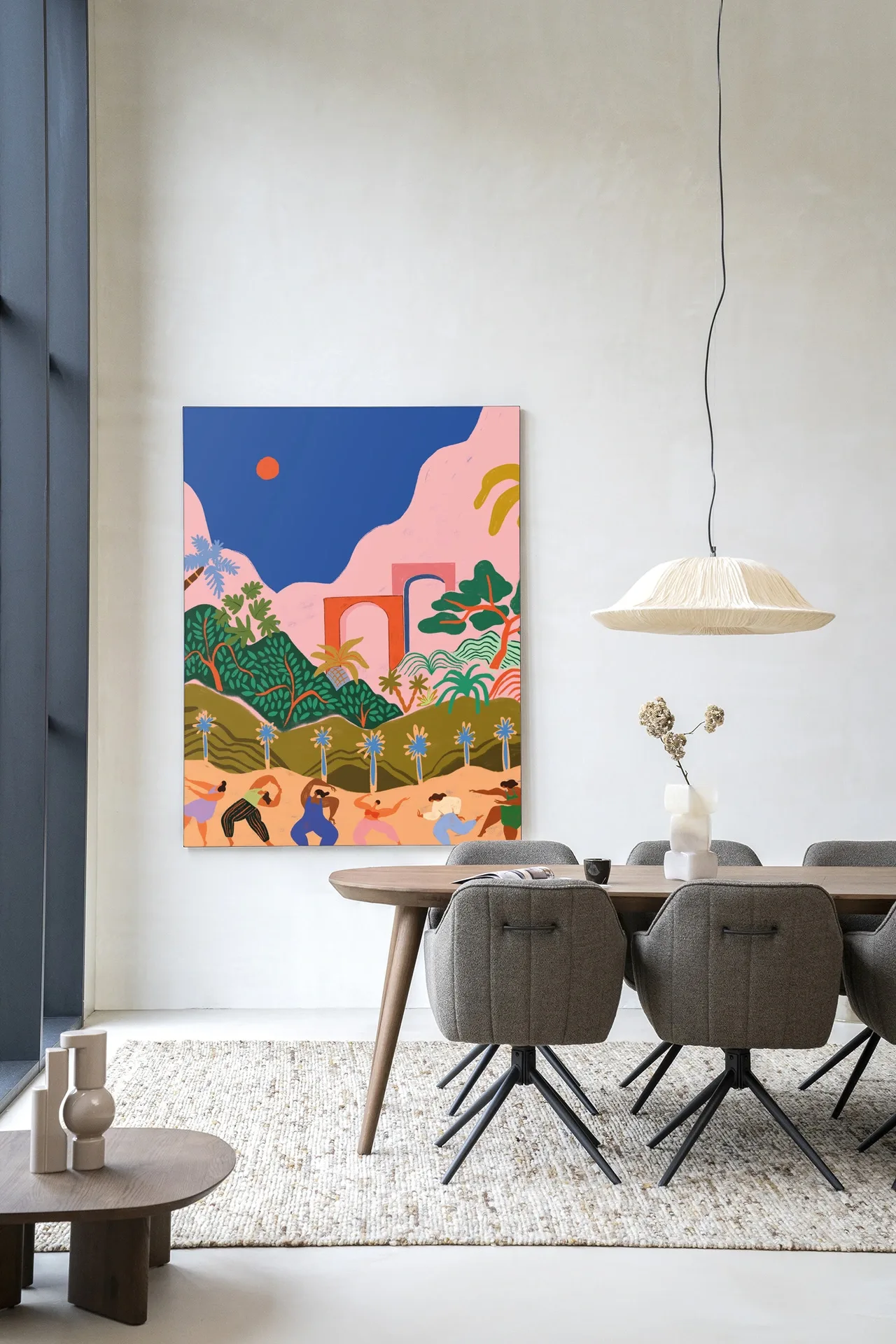 Een kleurrijk Wandkleed Dancing In Paradise - Urban Cotton hangt aan een witte muur boven een houten eettafel met grijze stoelen