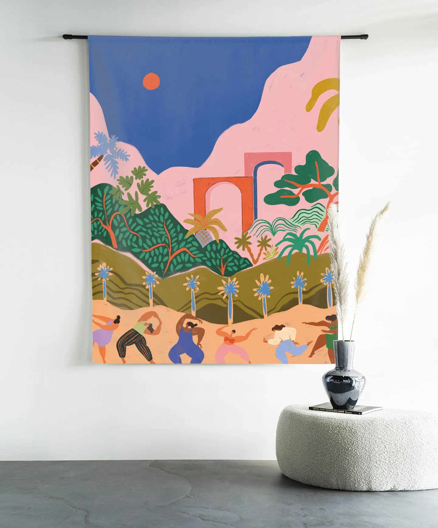 Een kleurrijk Wandkleed Dancing In Paradise - Urban Cotton hangt aan een witte muur boven een crèmekleurige poef met een vaas