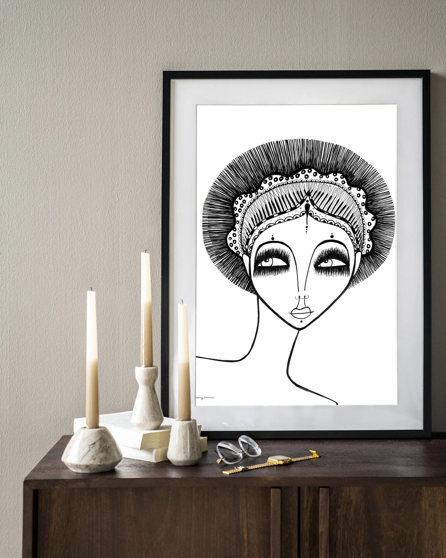 Een ingelijst Wandkleed Leyla - Urban Cotton hangt boven een houten dressoir met kaarsen en een bril
