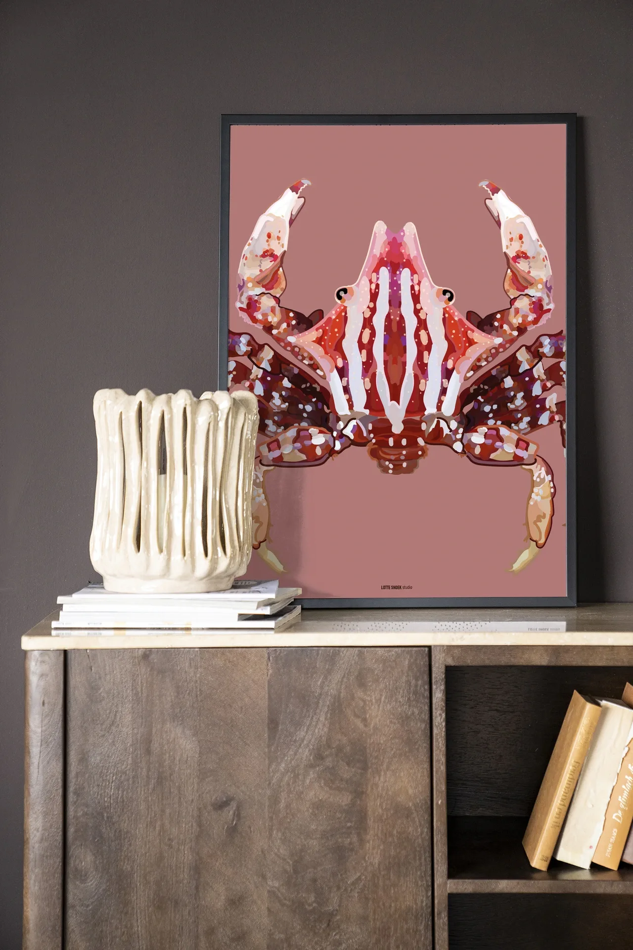 Wandkleed Villo Donus - Urban Cotton, ingelijst aan een donkere muur, met een vaas en boeken op een houten kast