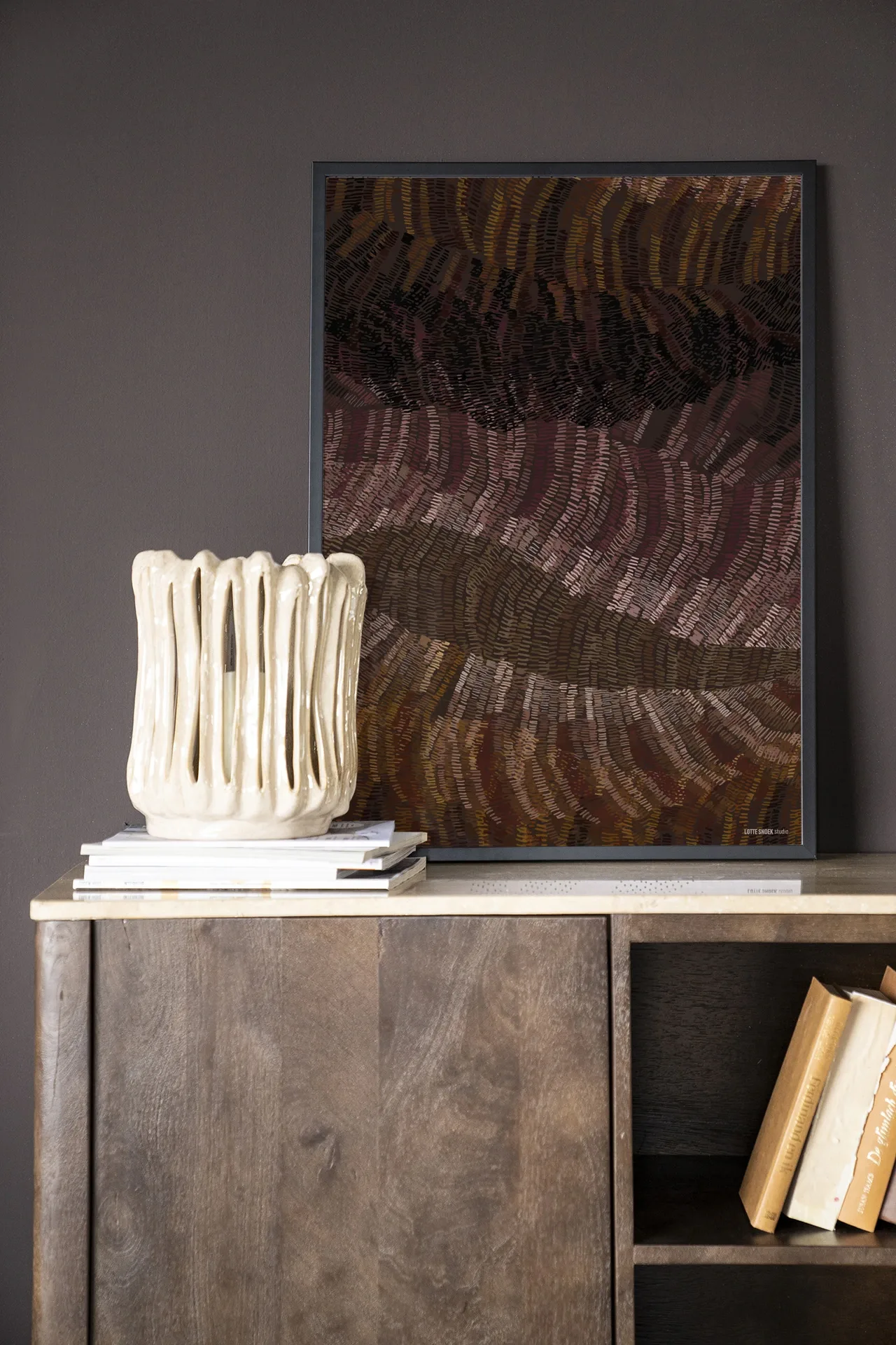 Het Wandkleed Olanan - Urban Cotton hangt aan een donkere muur boven een houten kast met boeken