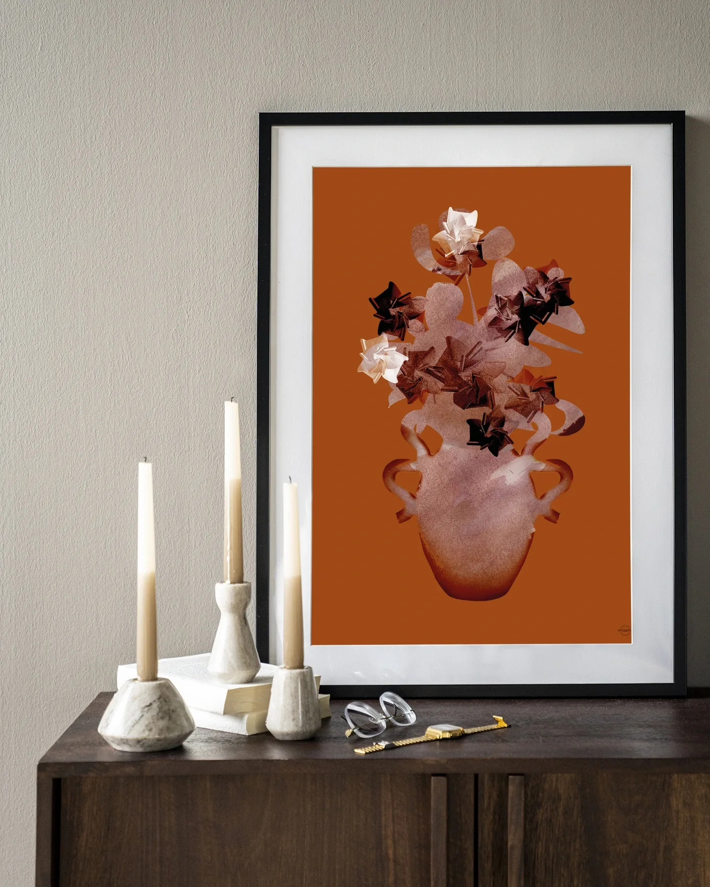 Een Art print Amphora Flowers - Urban Cotton in een zwarte lijst, staand op een donkerbruin dressoir met kaarsen en boeken