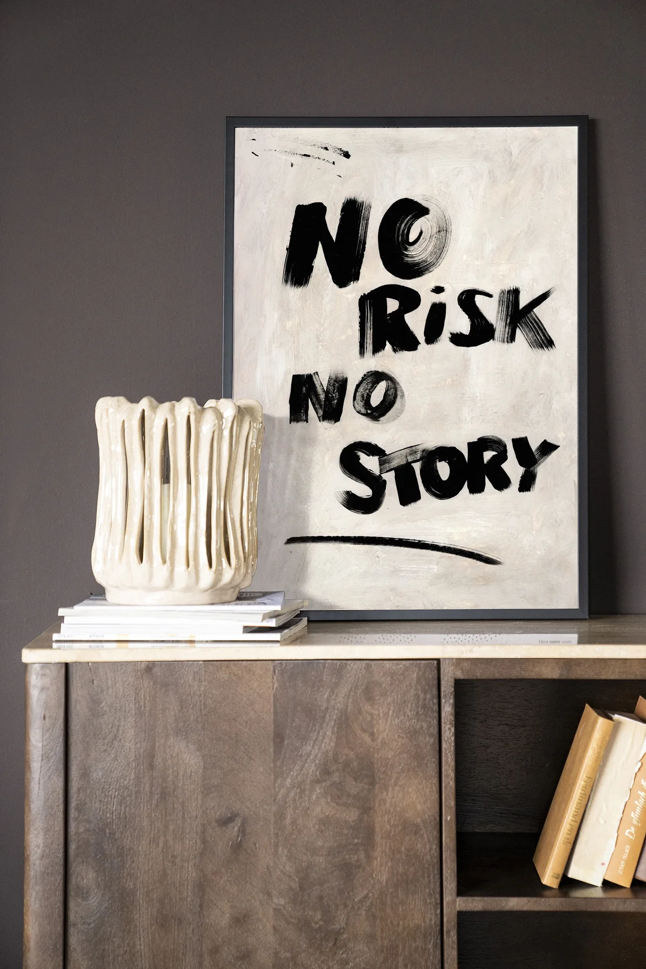 Wandkleed No Risk, No Story - Urban Cotton in een lijst, staand op een houten dressoir met boeken en een vaas