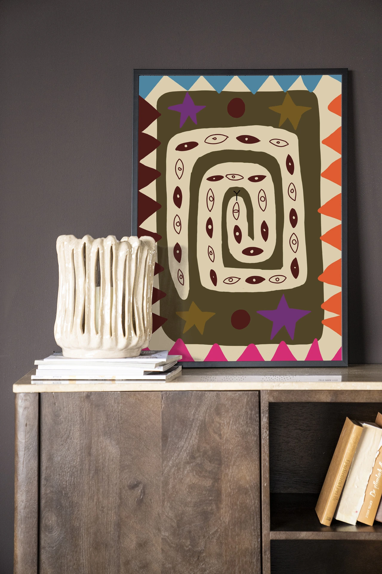 Wandkleed Snake Eye - Urban Cotton hangt boven een houten dressoir met boeken en een vaas