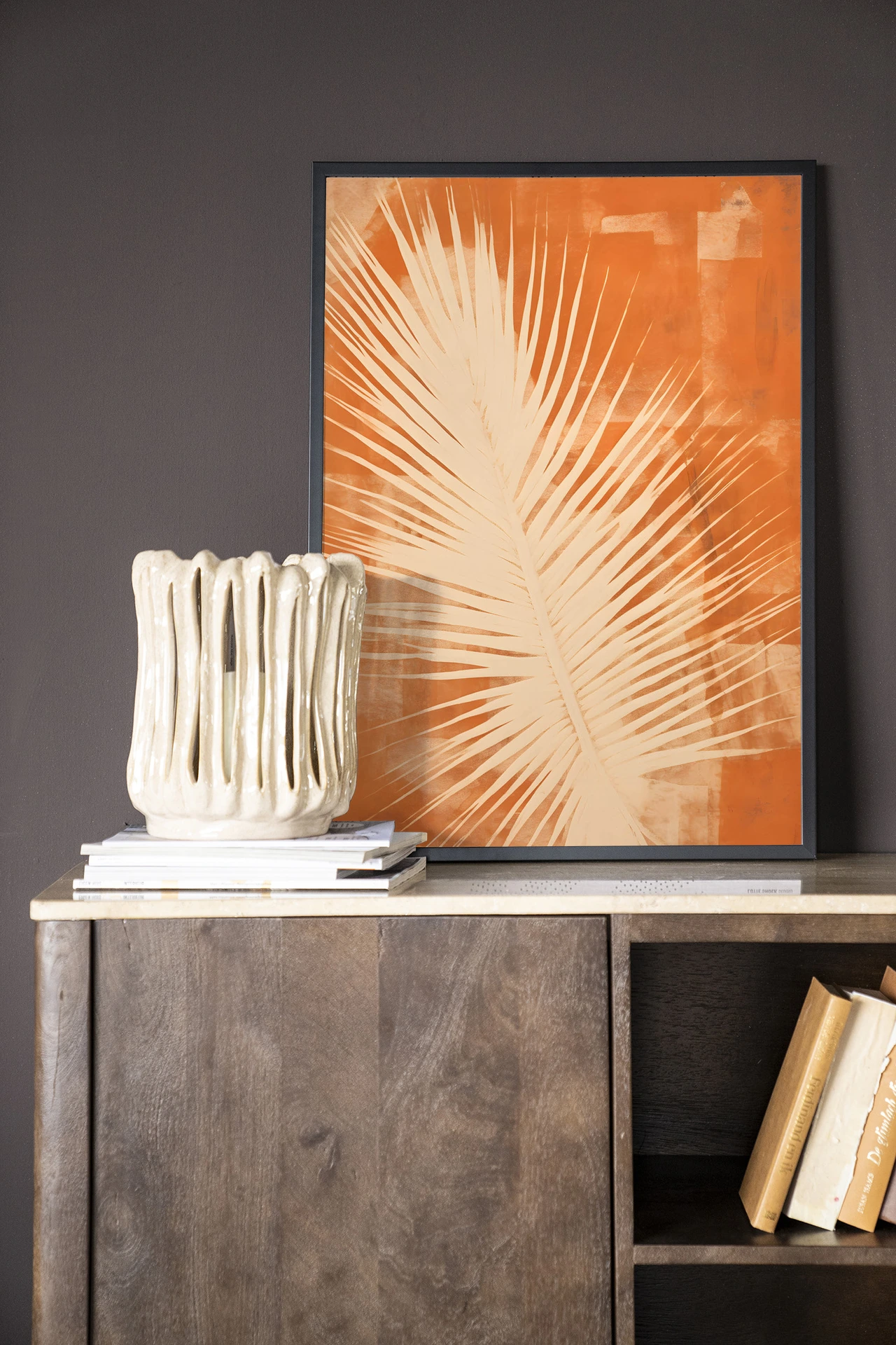 Het Wandkleed Rusty Leaf - Urban Cotton in een lijst, staand op een houten dressoir met boeken
