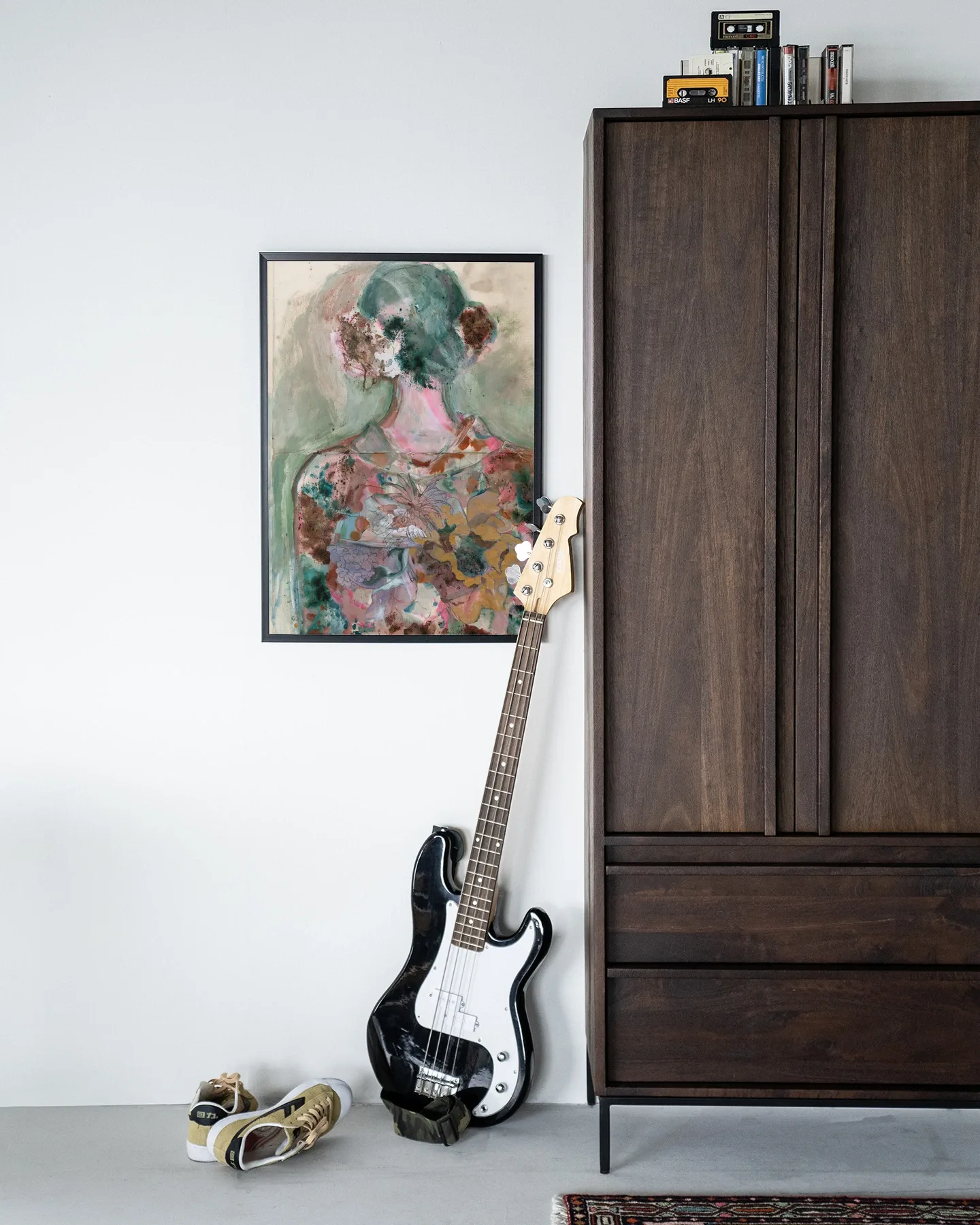Het Wandkleed Eclectic - Urban Cotton hangt aan een witte muur naast een donkerbruine kast, met een basgitaar en schoenen op de vloer