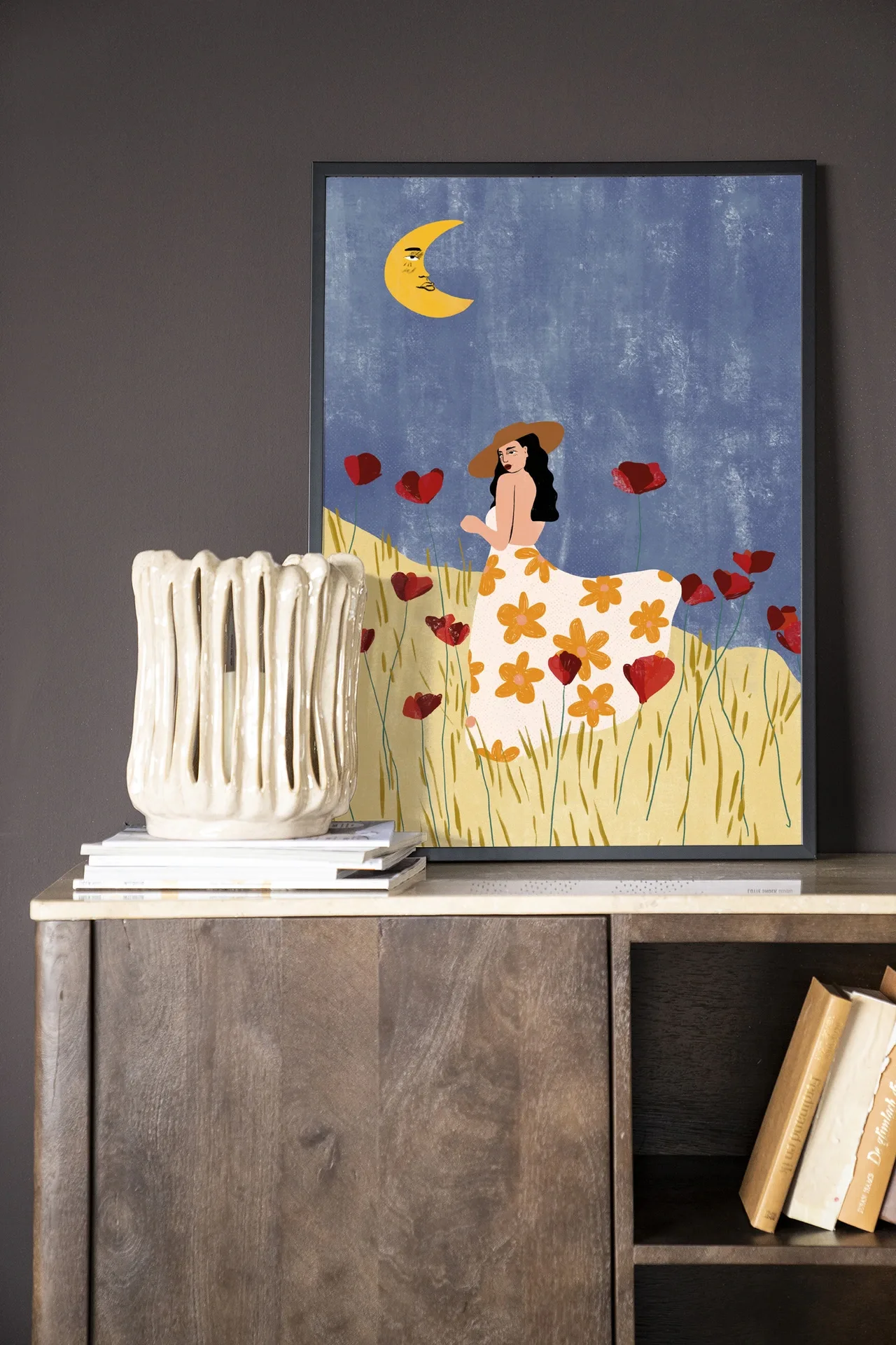 Wandteppich Girl In A Poppyfield - Urban Cotton hängt über einer Holzkommode mit einer Vase und Büchern
