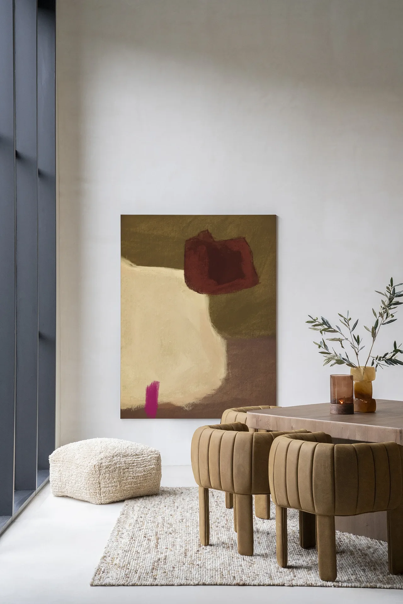 Wandteppich Navia - Urban Cotton hängt an einer weißen Wand über einem cremefarbenen Pouf und einem Holzesstisch mit Stühlen