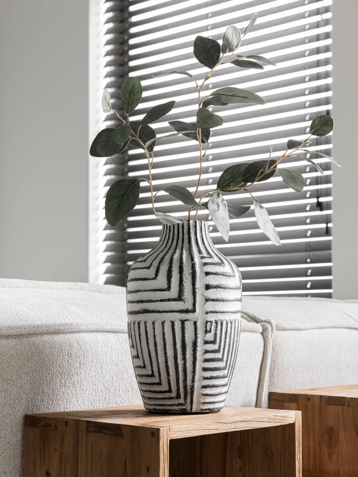Vase Braga mit einem grünen Zweig, stehend auf einem hölzernen Beistelltisch vor einem Sofa und Jalousien - MUST Living
