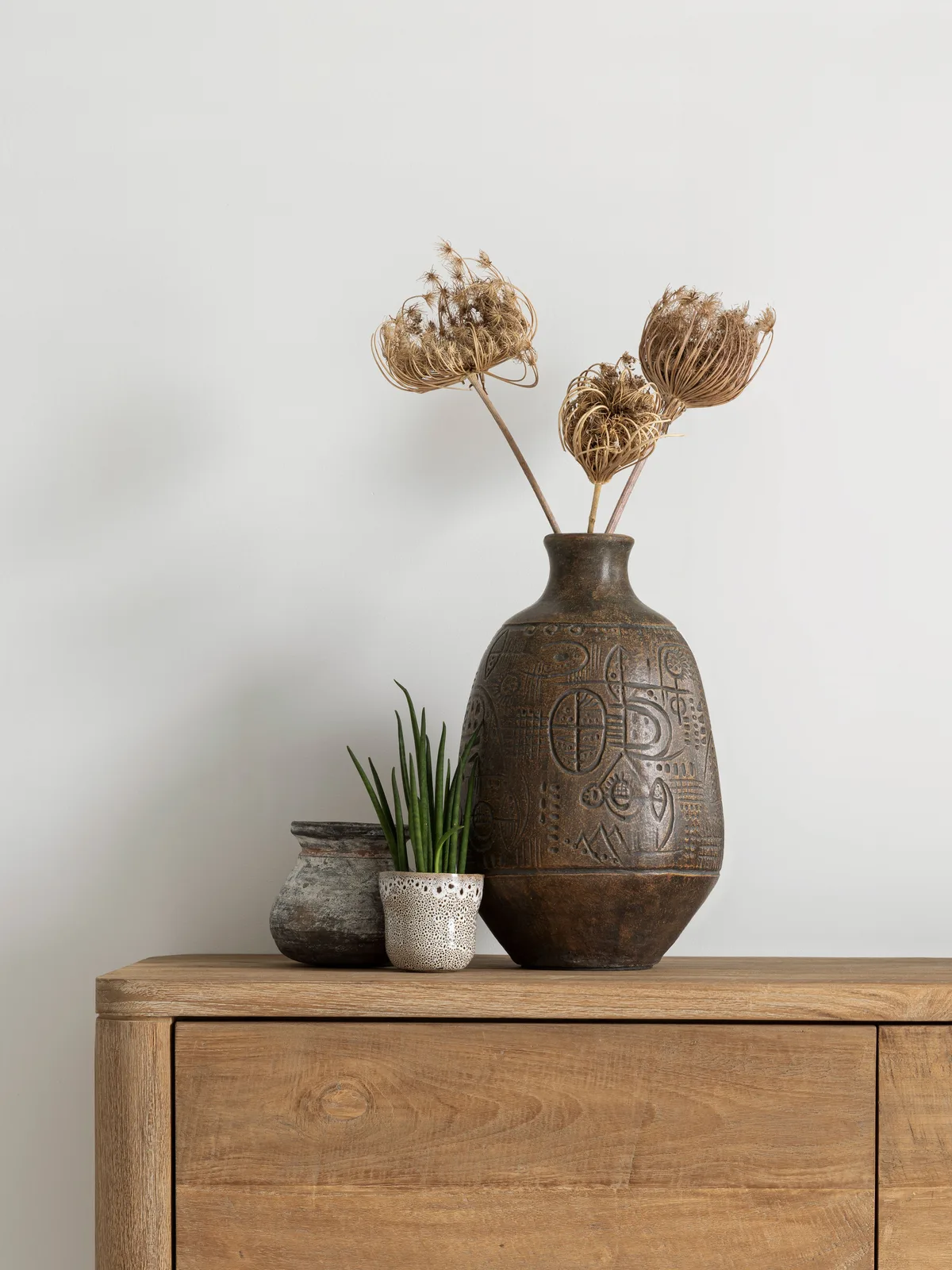 Die Vase Faro steht auf einem Sideboard aus Holz mit Trockenblumen und zwei kleineren Töpfen daneben - MUST Living