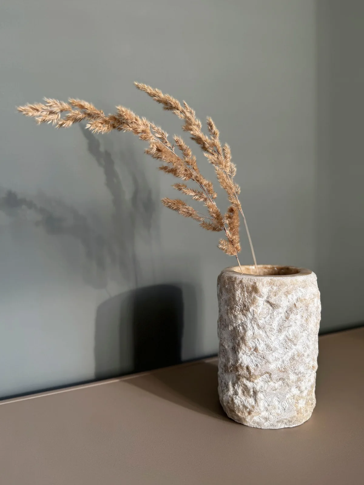 Vase Apollo mit getrockneten Gräsern auf braunem Untergrund, vor einer grünen Wand mit Schatten - MUST Living