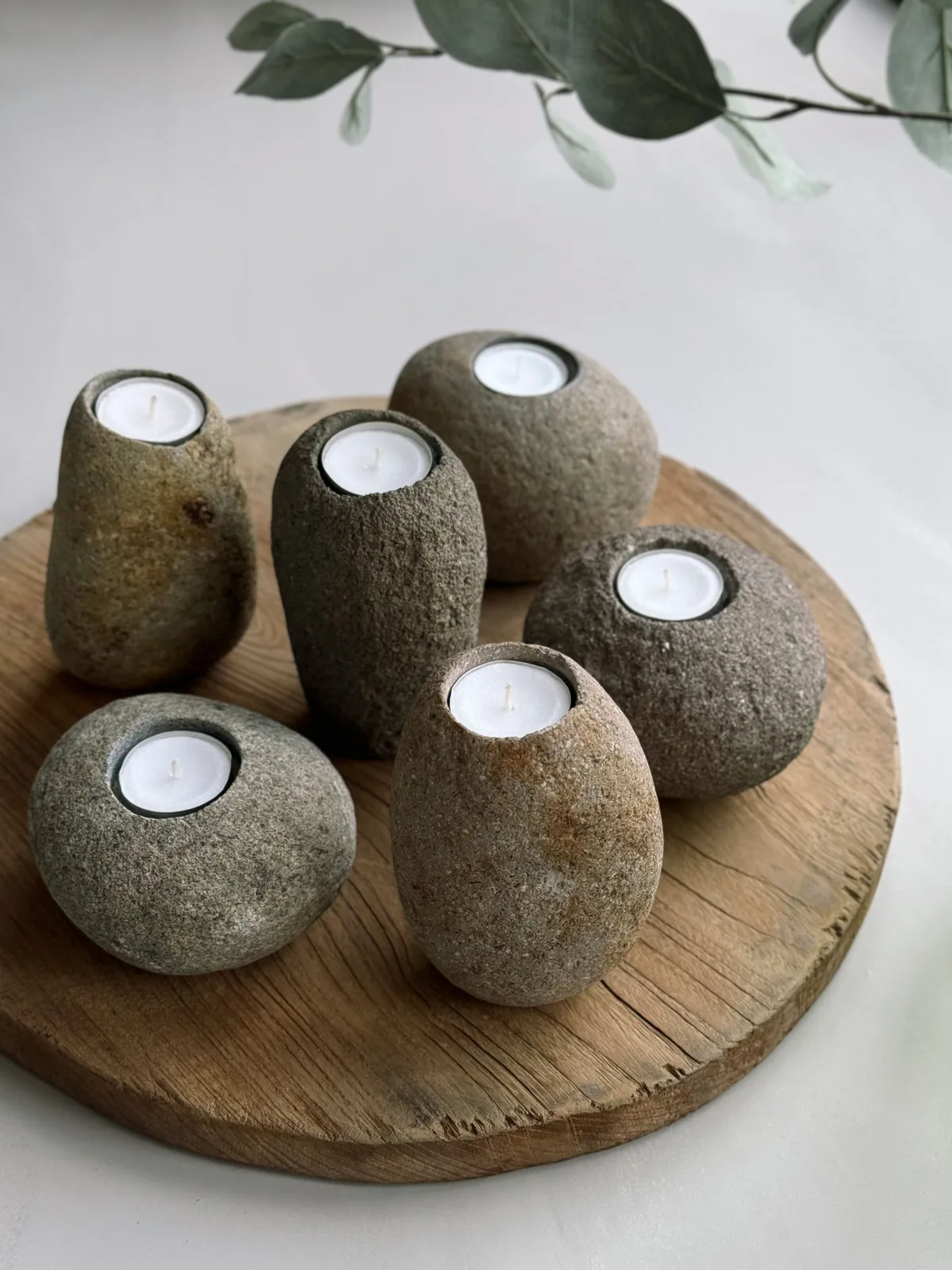 Sechs Kerzenhalter Riverstone hoch mit brennenden Teelichtern auf einer Holzscheibe mit einem grünen Zweig im Hintergrund - MUST Living