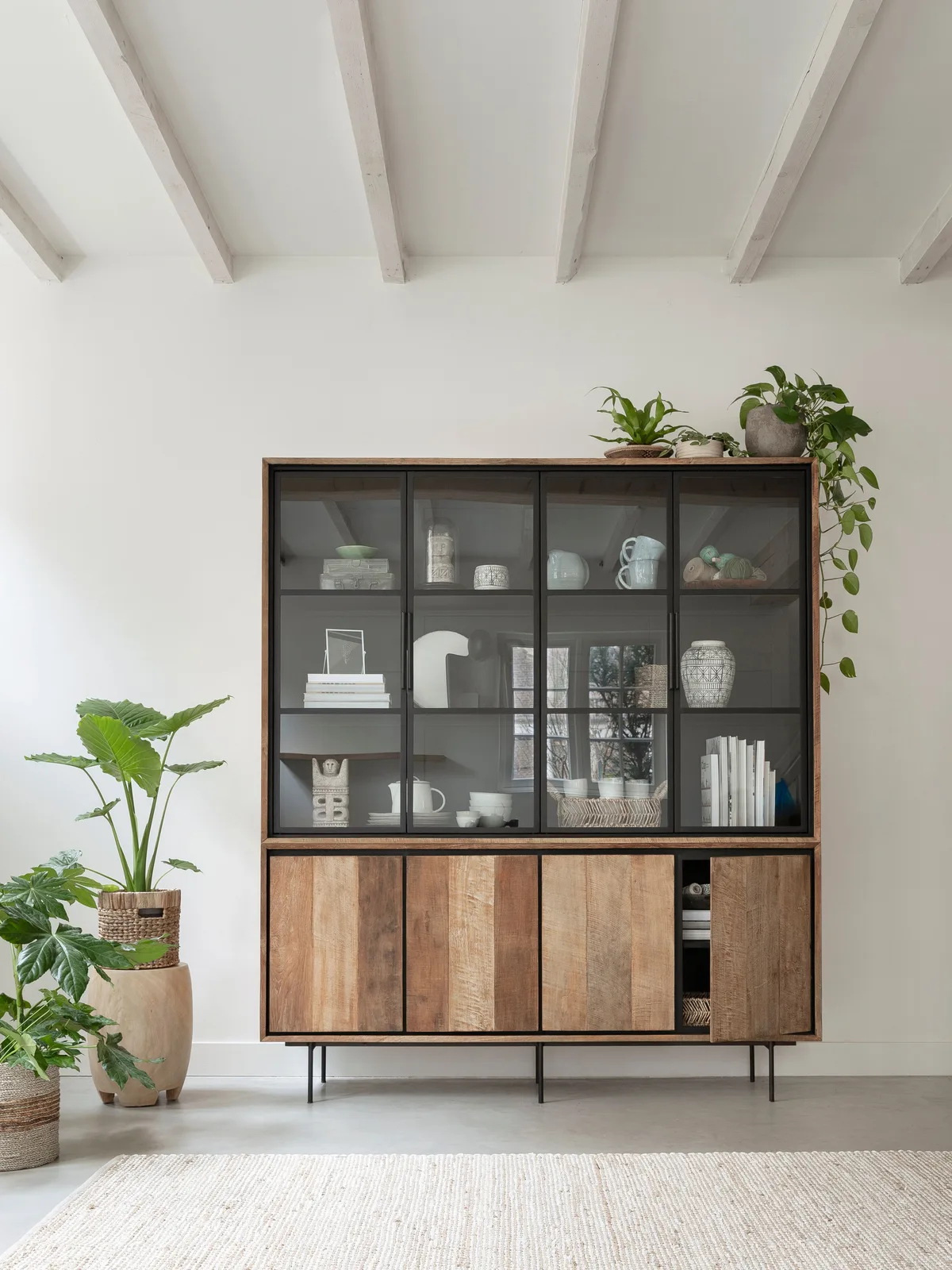 Der Vitrinenschrank Metropole in einem hellen Wohnzimmer mit Pflanzen und einem cremefarbenen Teppich - DTP Home