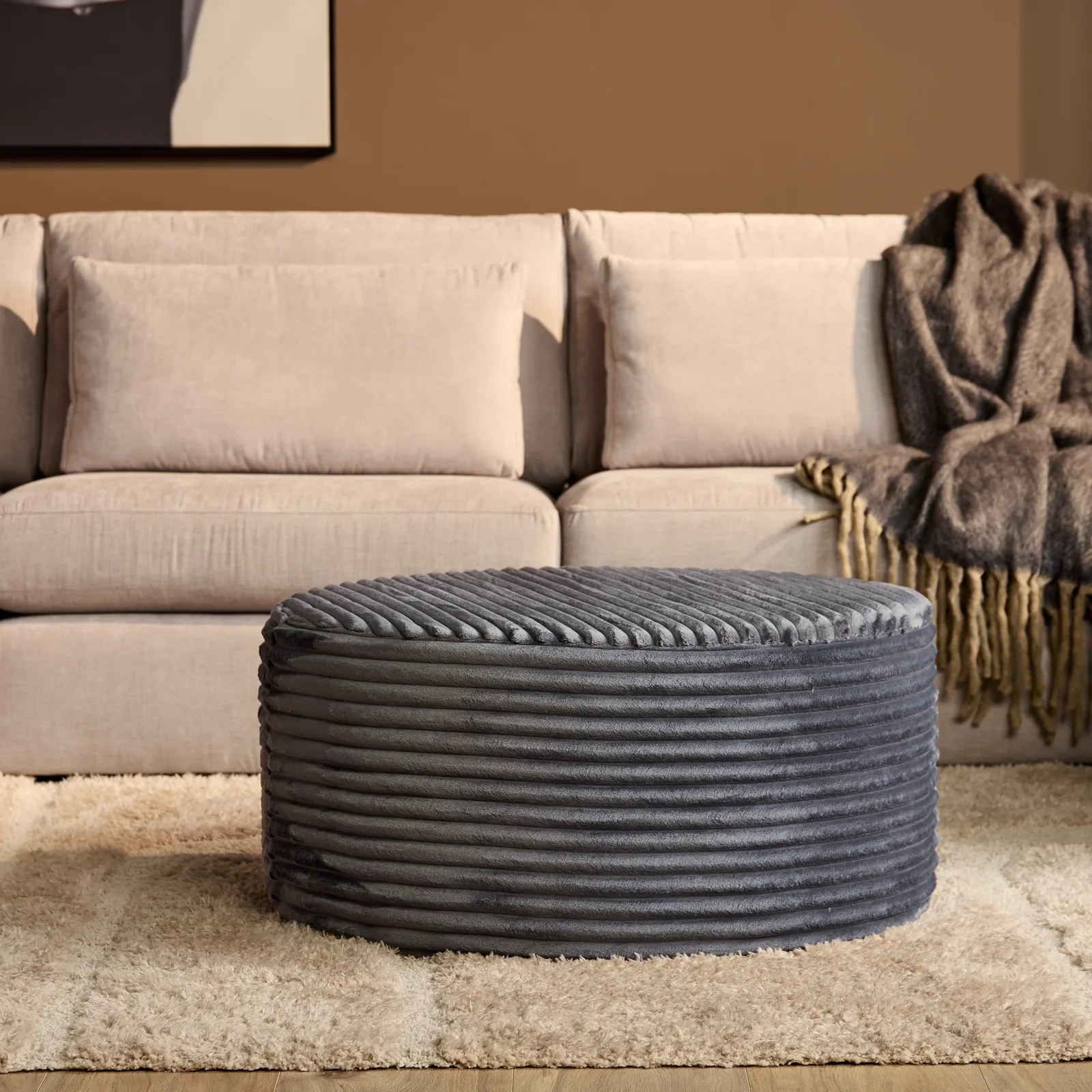 MySofa Runder Hocker in Dunkelgrau mit Rippenstoff, auf einem hellen Teppich vor einem Sofa stehend
