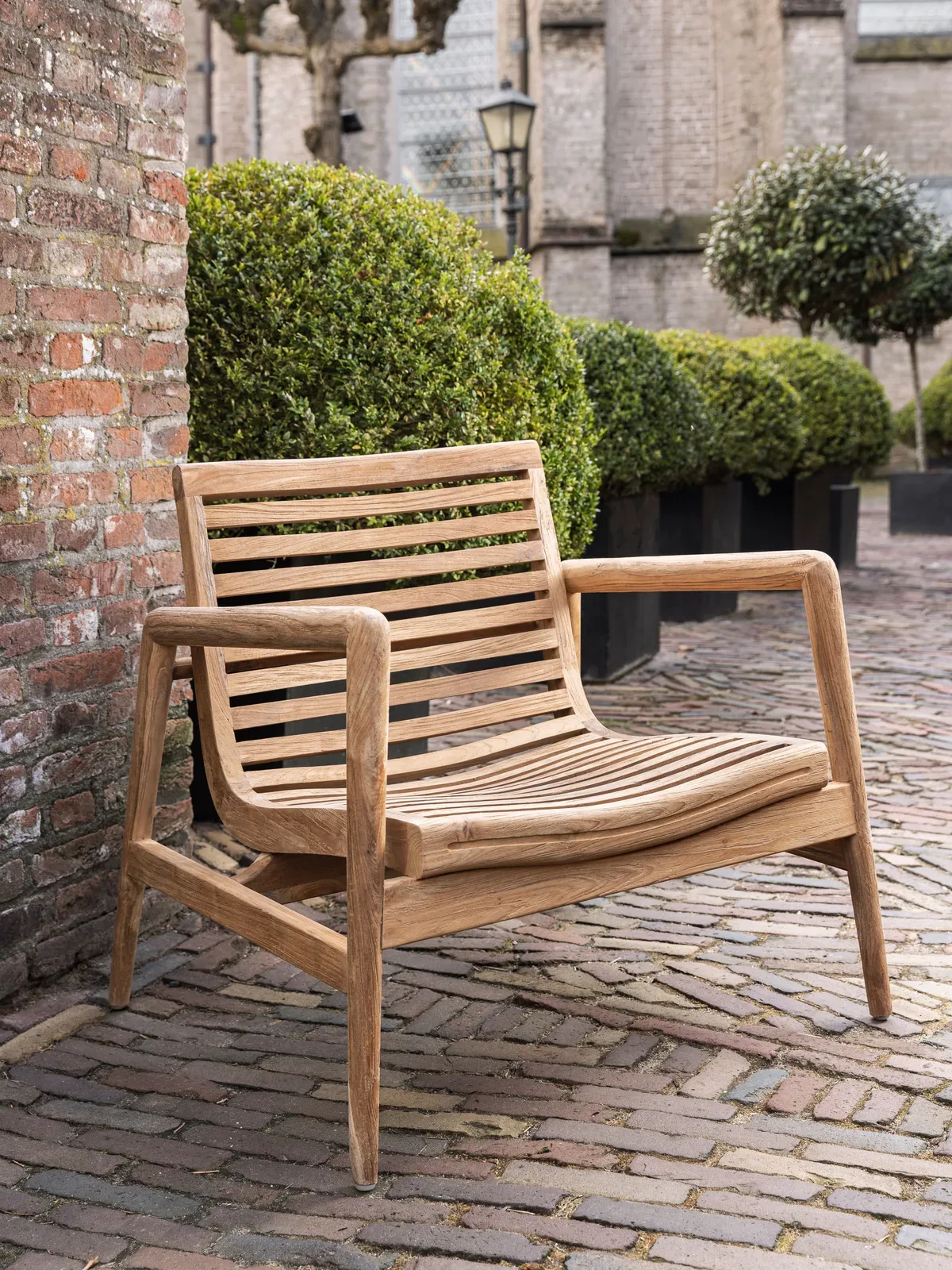 Loungestoel Ray auf einer Steinterrasse neben einer Ziegelmauer und grünen Sträuchern - MUST Living
