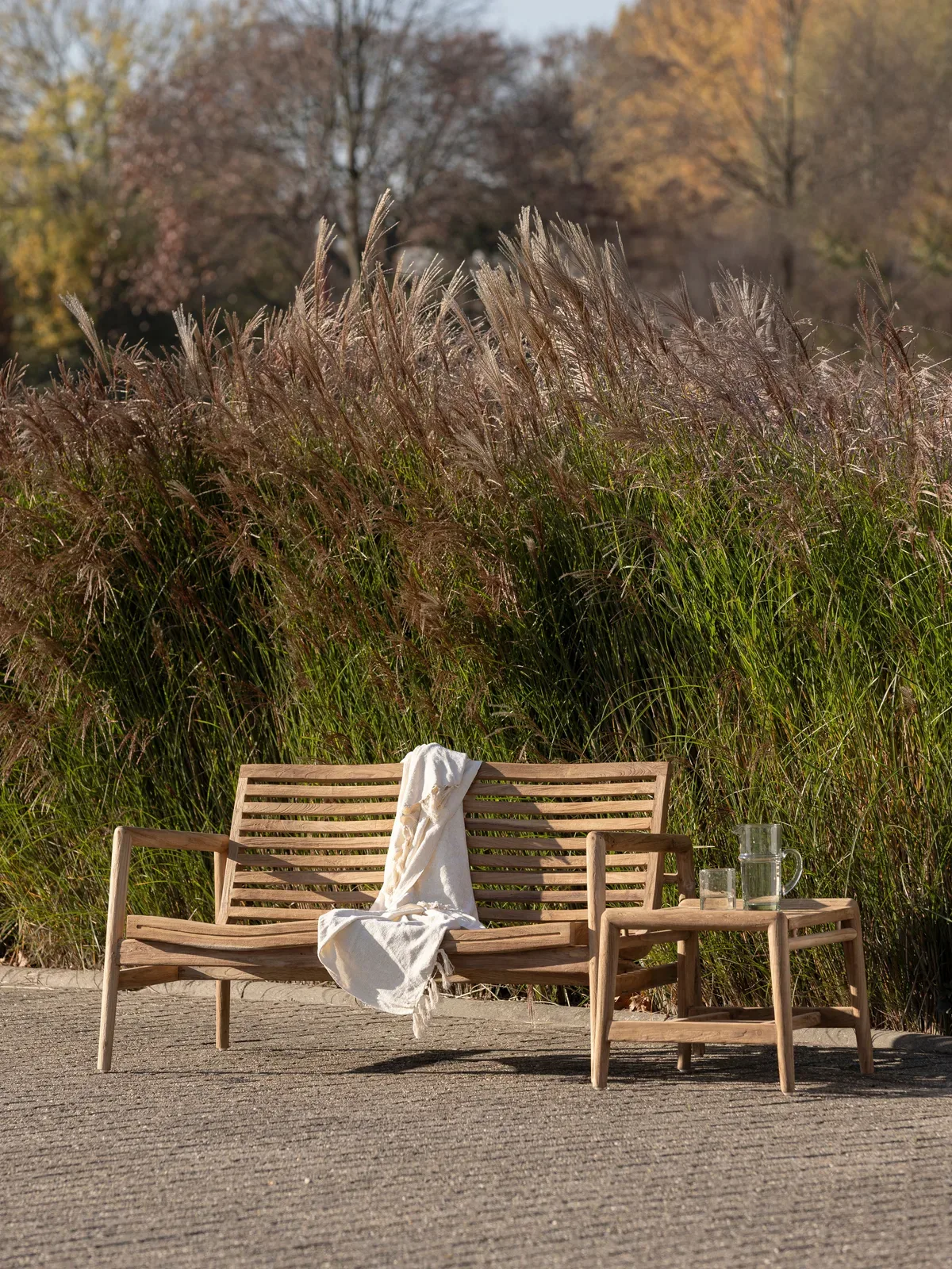 Gartenbank Ray 2-Sitzer mit einem Plaid und Beistelltisch in einem grünen Garten mit hohem Ziergras - MUST Living