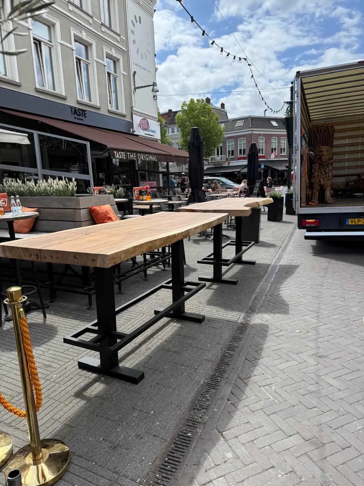 Ein Bartisch aus Suarholz Baumstamm mit schwarzem Metallgestell steht auf einer Terrasse zwischen anderen Tischen - World of Brosi