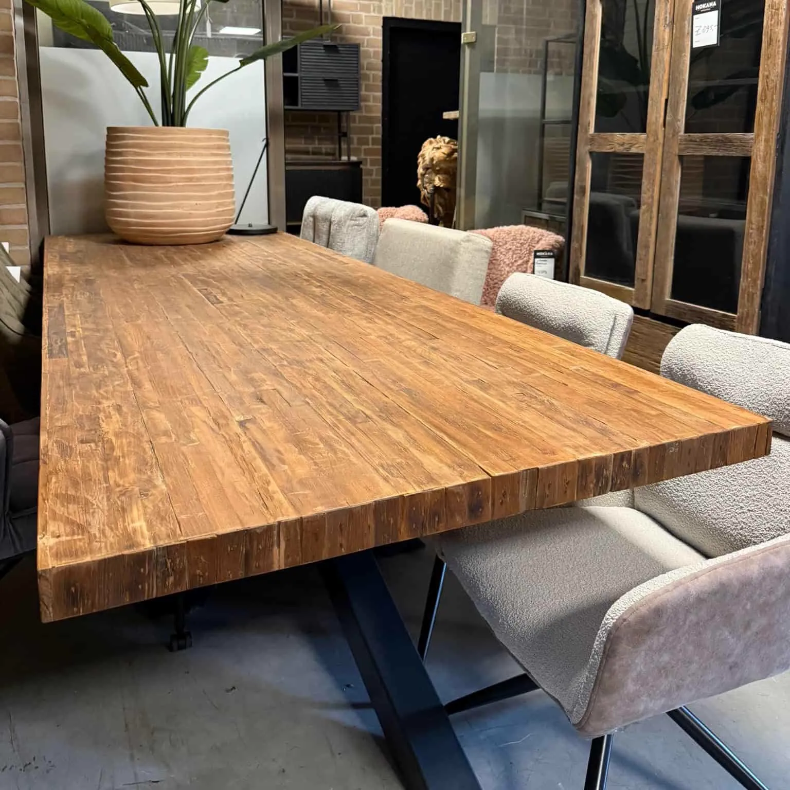 Der Donny Teakholz-Esstisch Massiv 280 cm steht im Mittelpunkt eines stimmungsvollen Showrooms, umgeben von Stühlen - World of Brosi