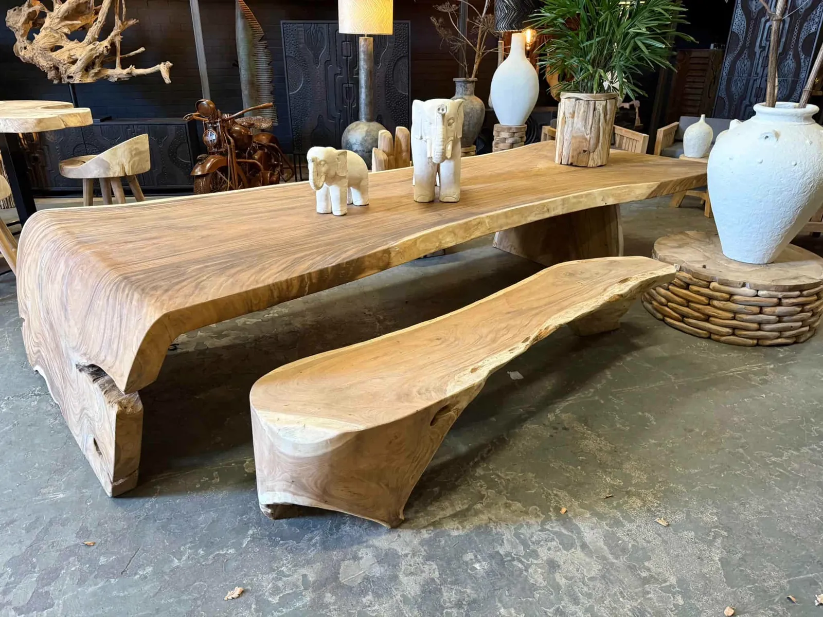 Wasserfall Baumstammtisch Suarholz 440cm mit passender Bank, dekoriert mit weißen Elefanten und Vasen in einem stimmungsvollen Showroom - World of Brosi