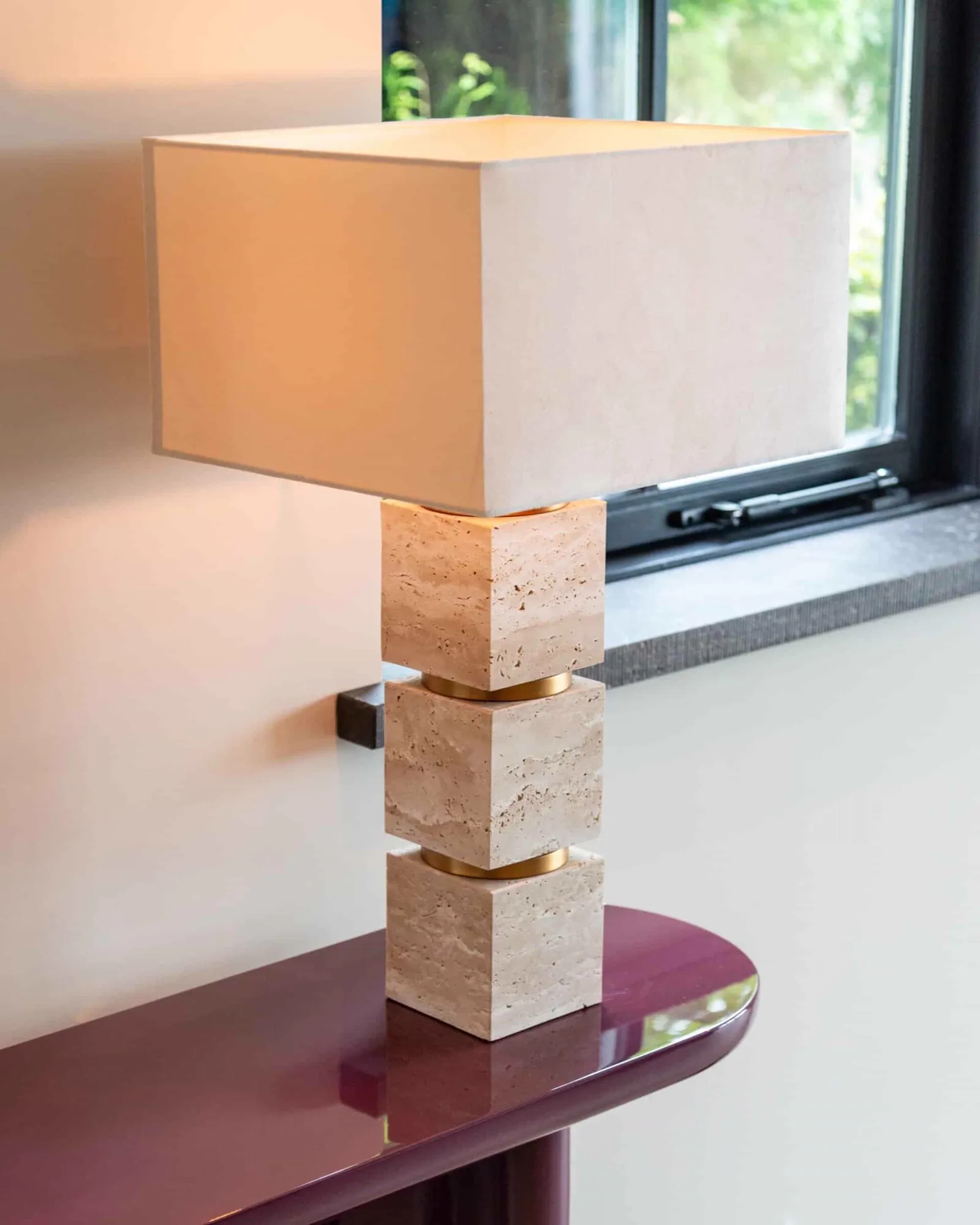 Tischleuchte Jaella beige auf einem lila Beistelltisch, mit einem Fenster und Grün im Hintergrund - Richmond Interiors