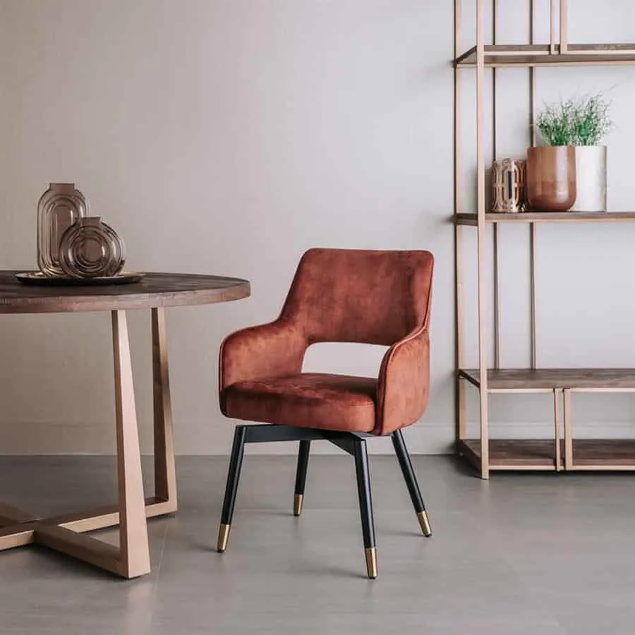 Lenno Sessel aus Stoff Adore 24 Copper, drehbar, mit schwarzen Beinen und goldenen Akzenten, an einem runden Tisch und einem offenen Regal - Tower Living