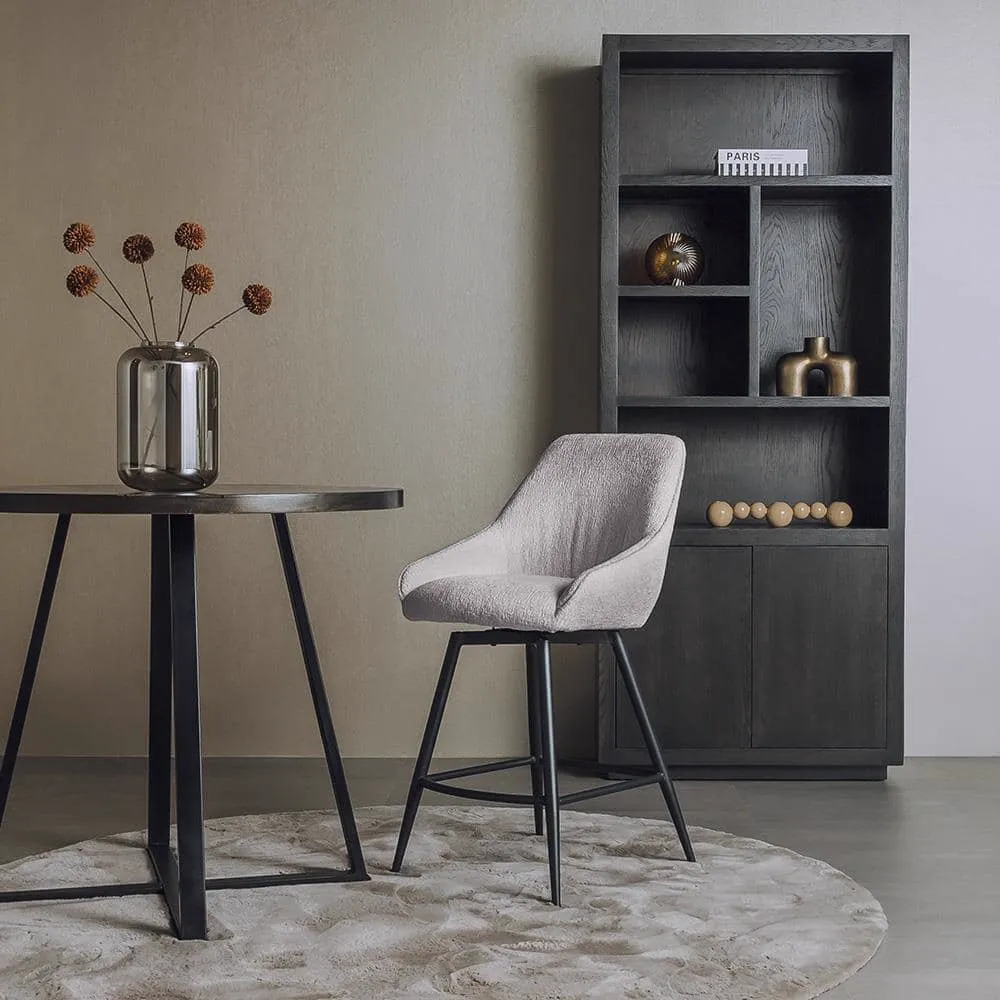 De Trino Barstoel - fabric Trenza 08 oyster (draaibaar) staat in een moderne setting naast een hoge tafel en boekenkast - Tower Living