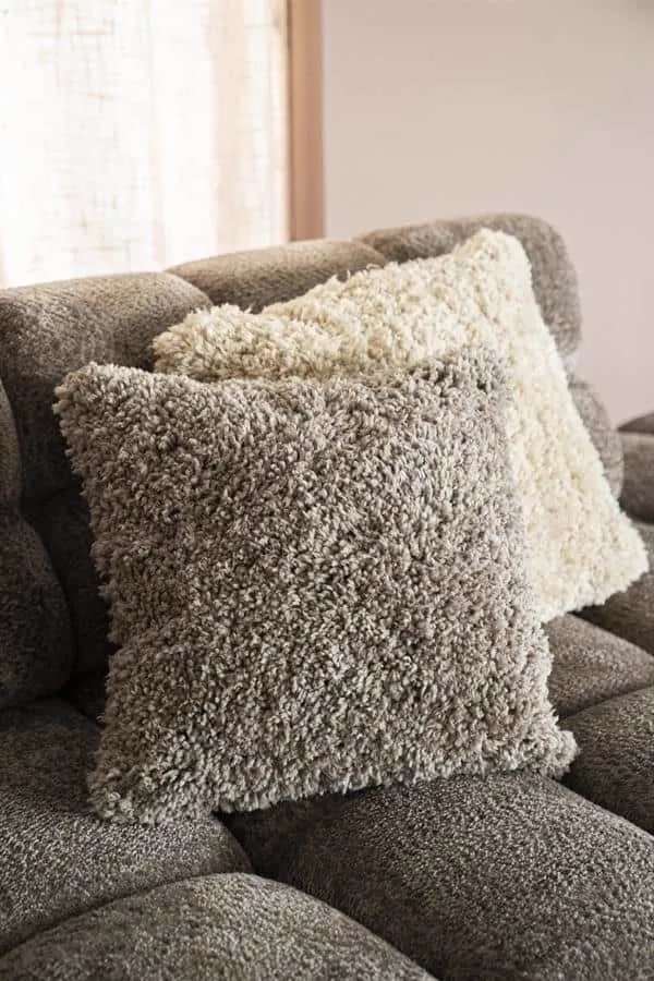 Zwei zottelige Kissen, ein taupefarbenes Kissen Fez taupe und ein cremefarbenes, liegen auf einem Sofa - By-Boo