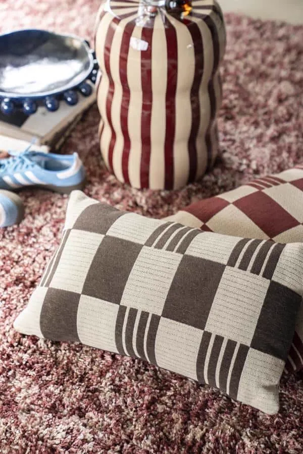 Das Kissen Twist braun liegt auf einem flauschigen Teppich, mit weiteren Wohnaccessoires im Hintergrund - By-Boo