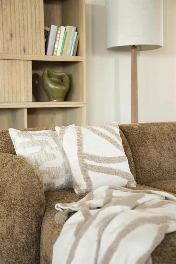 Kissen Majani Taupe mit abstraktem Muster auf einem braunen Sofa, neben einem Holzregal und einer Lampe - By-Boo