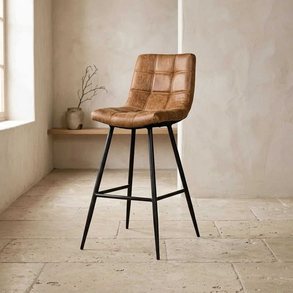 De Navarra Barstoel in cognac stof met zwarte poten staat in een lichte studio met een raam en een houten plank - Tower Living
